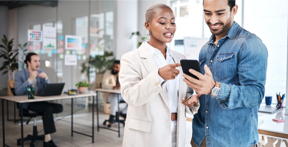 A man in a jacket holds a smartphone while a businesswoman points to the screen.