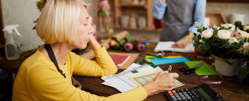 A woman sits at a desk with a calculator, surrounded by a vase of flowers, focused on her work.