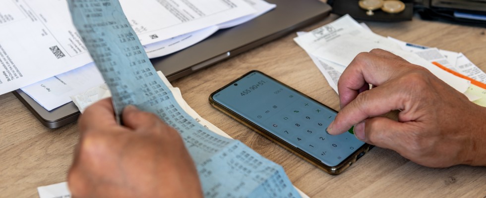 A man holds a receipt in one hand while typing on a calculator with his other. Stacks of receipts and other financial documents sit on a desk.