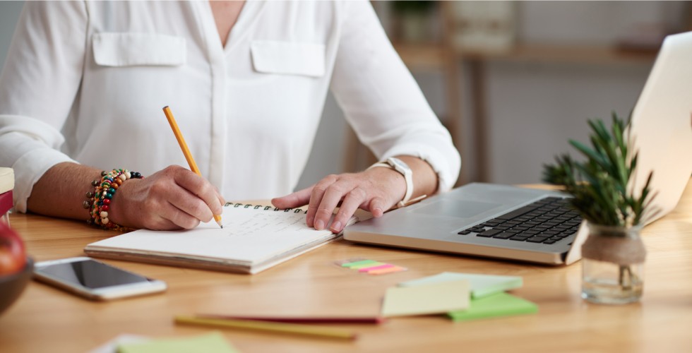 A woman sits at a desk, writing in a notebook, focused on her task with a pen in hand.