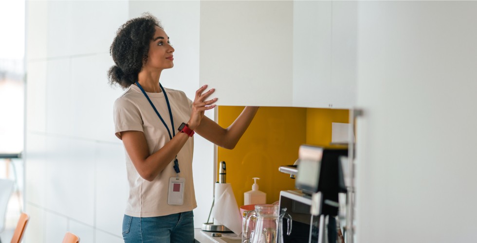 A woman is positioned before an office kitchen cabinet, emphasizing the neat arrangement and contemporary aesthetics of the kitchen.
