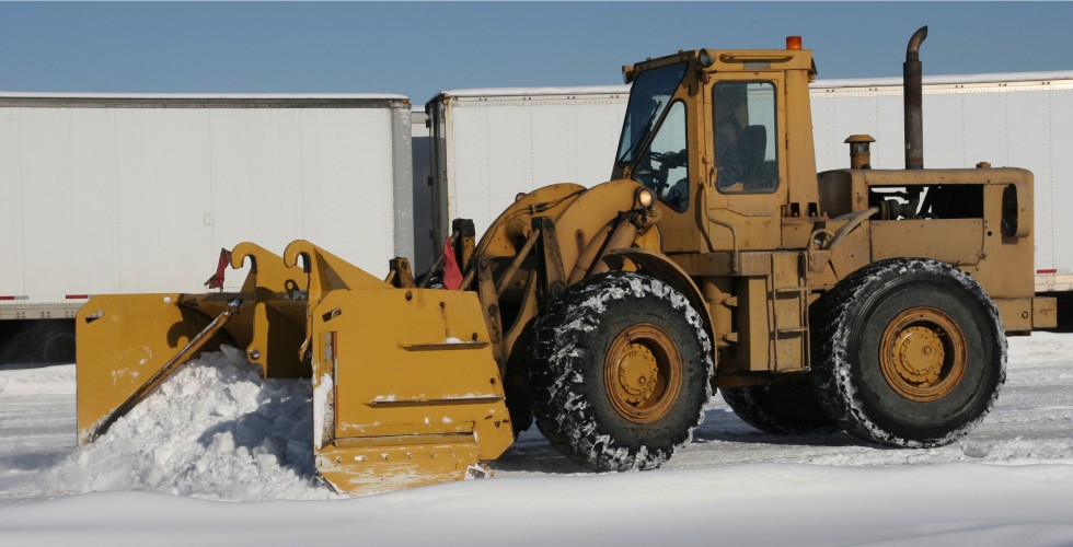A bright yellow bulldozer uses its large shovel to remove snow from a parking area.