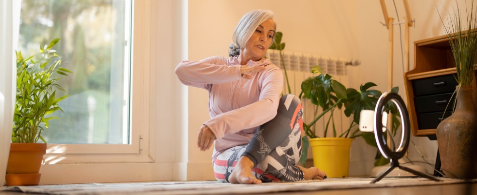 A senior woman in athletic wear doing a yoga stretch at home, with a camera on a tripod recording her.