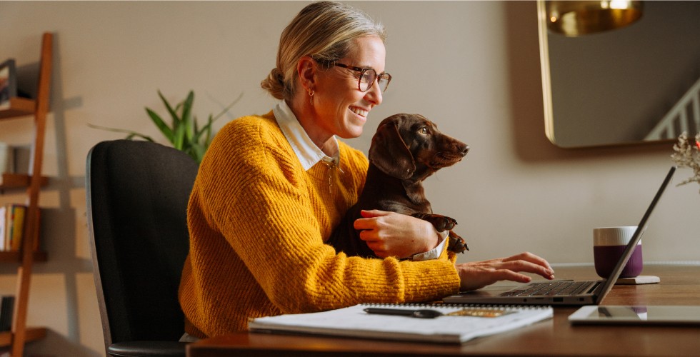 A woman working from home holds a small dog in her lap while typing on her laptop. Her desk has a coffee mug and paper notepad resting on it.