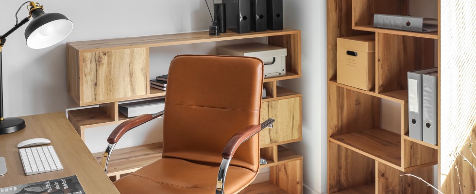 A pristine home office space has a variety of shelves, storage binders, boxes, a work desk, table lamp, and leather chair.