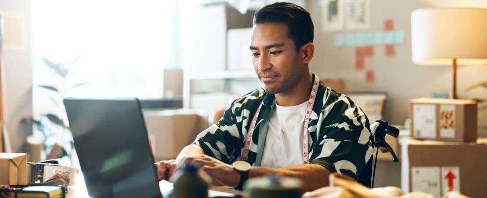 A man working from home types away on his laptop. He has a tape measure draped around his neck and shipping boxes stacked behind him.