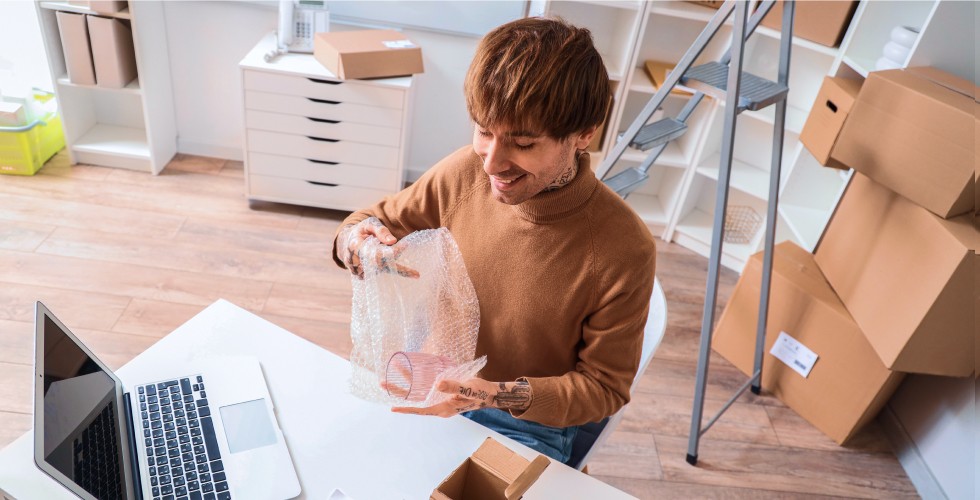 A man sitting at a work desk with a ladder and storage boxes behind him unwraps a drinking glass.