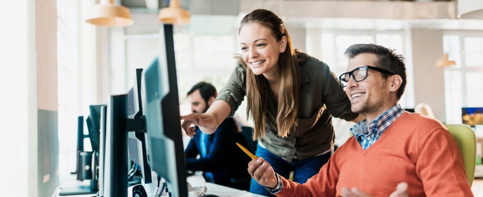 Two coworkers smile as one of them points to a computer screen in front of them.