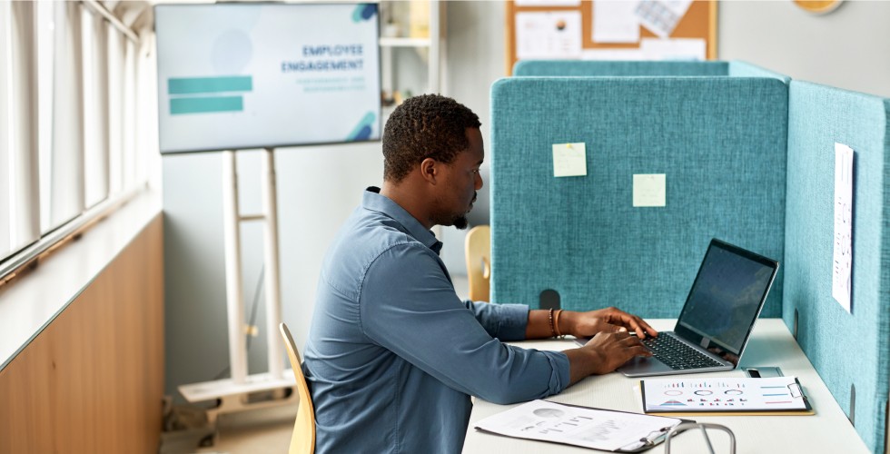 A man works in an office cubicle, where the walls are low and covered with teal colored fabric.