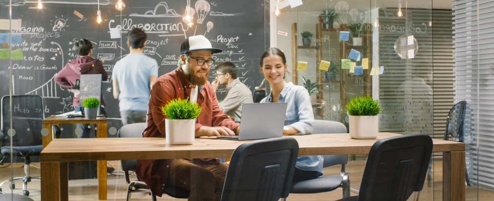 A casual atmosphere workspace has coworkers sitting around a desk and writing on a blackboard in the background.