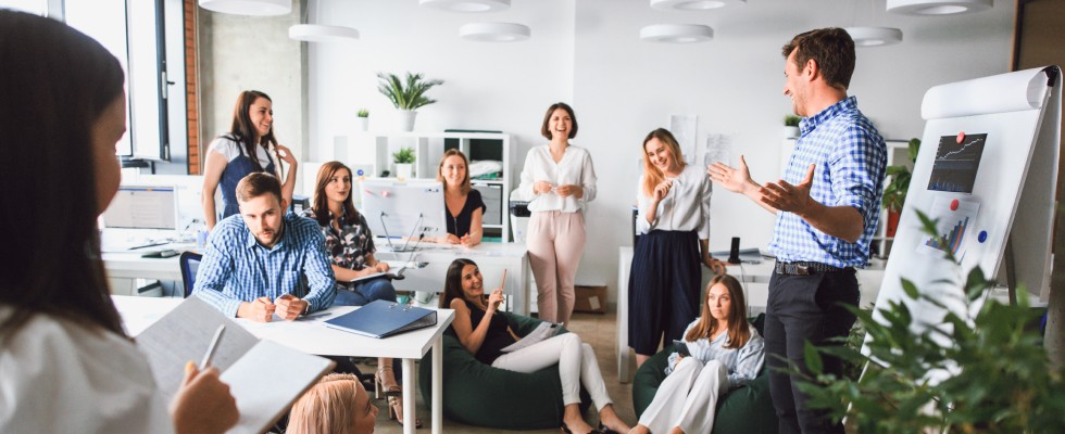 A business professional gives a presentation to a room full of coworkers who are smiling and taking notes.