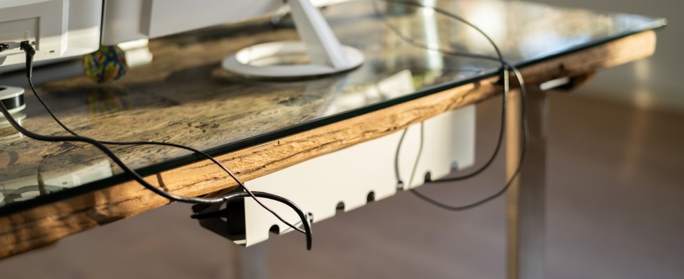 A close-up of a glass-top desk with computer monitor cables organized underneath. The image focuses on the minimalist and organized setup of the workspace.