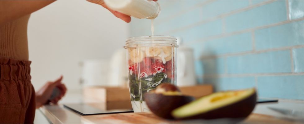 Hands pouring a small bottle of milk into a blender cup filled with cut bananas, strawberries, and spinach.