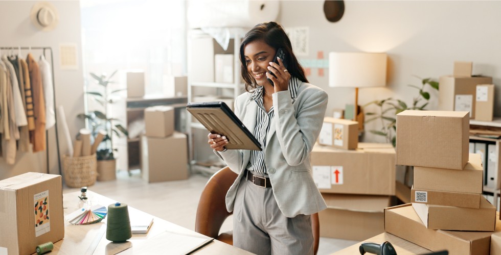 A woman in a business suit speaks on her cell phone while seated in a room filled with unpacked boxes.