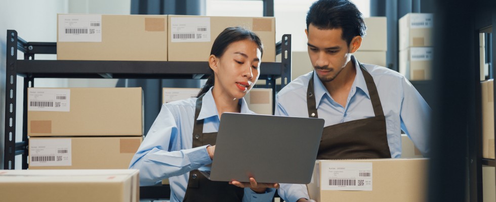 Two individuals collaborating in a warehouse, surrounded by stacked boxes, efficiently organizing inventory.