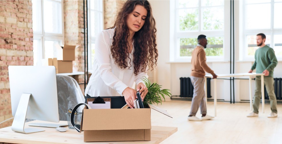 A woman is packing a cardboard box in a modern office setting, preparing for a move or organization task.