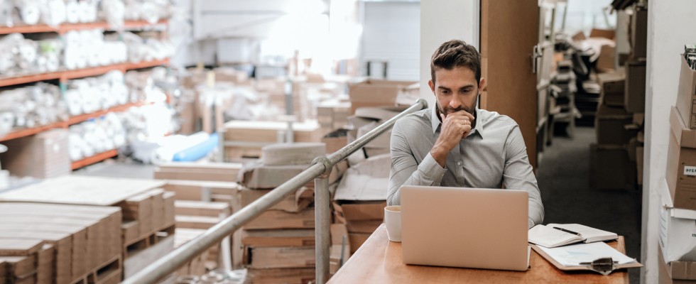 A man sits at a table with a laptop in a spacious warehouse, surrounded by shelves and industrial equipment.