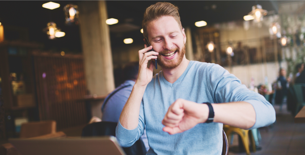 A man in a restaurant smiles as he checks his watch while taking a phone call.