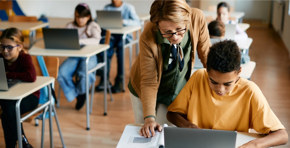 A teacher assists a student with a laptop, fostering a supportive learning environment.
