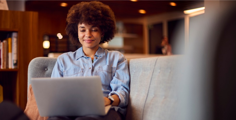 A young woman with curly hair, smiling as she works on her laptop while sitting on a comfortable sofa in a cozy, modern setting.