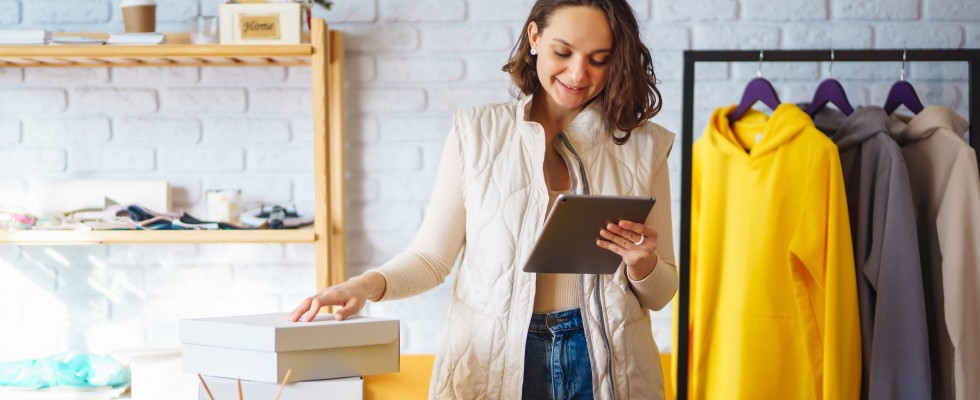 A woman wearing a beige vest, standing in a brightly lit store with clothing racks and shelves, happily using a tablet while working.