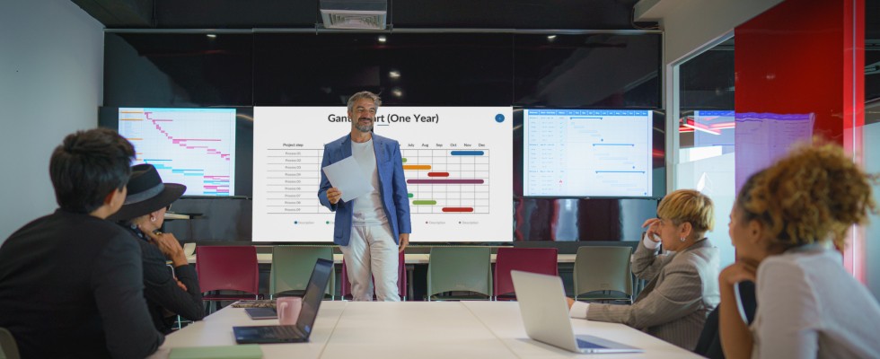 A businessman holding a paper, presenting data on large screens behind him to a group of people seated around a conference table.