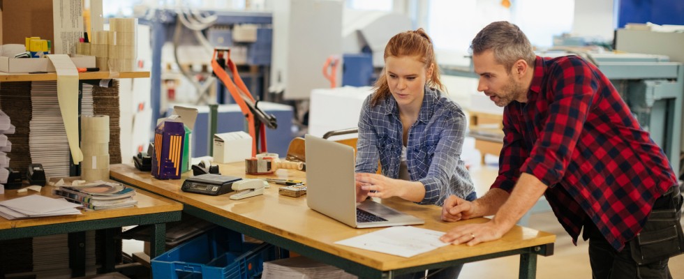 Two individuals collaborating on a laptop inside a factory setting, focused on their work amidst industrial surroundings.
