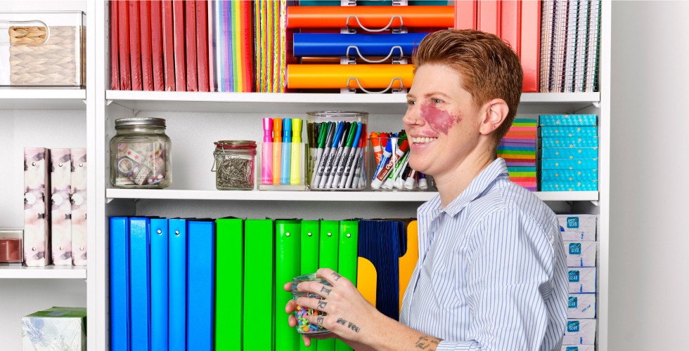 A person with bright paint on their face organizing colorful binders, folders, and stationery supplies on a shelf in an office or classroom.