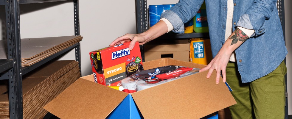 A person packing Hefty trash bags and other cleaning supplies into a large cardboard box in a storage room.