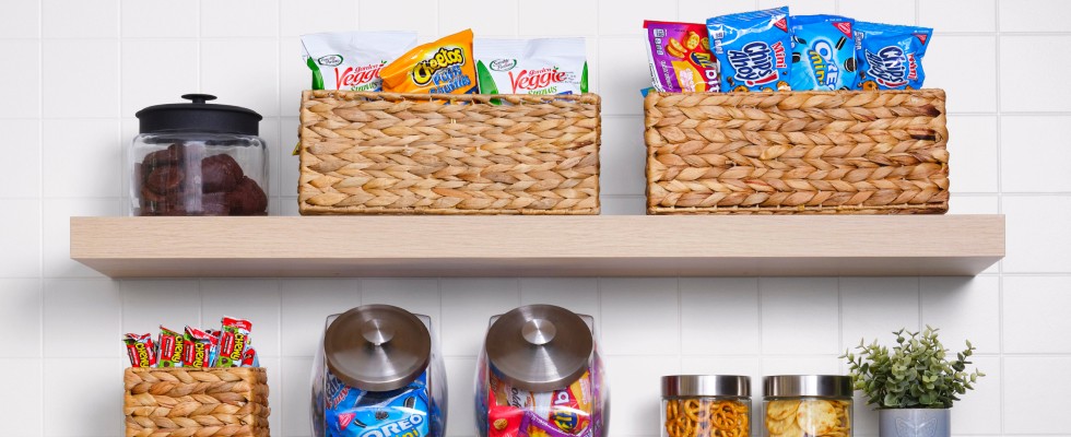 A shelf in a kitchen stocked with various snacks and packaged foods in jars and woven baskets.