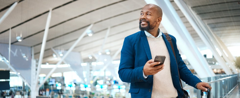A man in a suit stands with his luggage inside an airport terminal, holding out his smartphone.