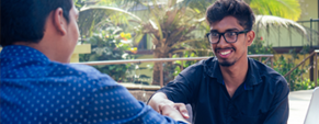 In an outdoor setting, a smiling, bespectacled, male student shakes hands with a male interviewer.
