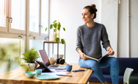 A woman sits on the edge of a desk, a book in her lap. The desk features a laptop, tablet, and various decor.