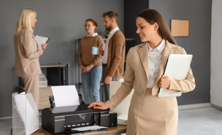 A woman examines a printer while holding a laptop. Professionals gather in the background of a clean office space.