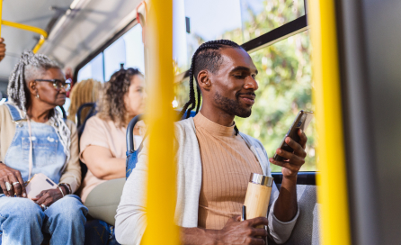 A man riding a bus on a sunny day smiles while looking at his smartphone.