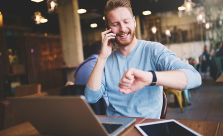 A man in a blue shirt sits in a busy office and takes a phone call while looking at his watch.