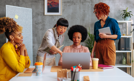 A diverse group of colleagues discuss a topic around a laptop in an office setting.  An individual is seen pointing toward the laptop as the others observe.