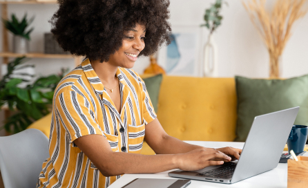 A woman smiles as she types on a laptop in a bright and cozy environment.