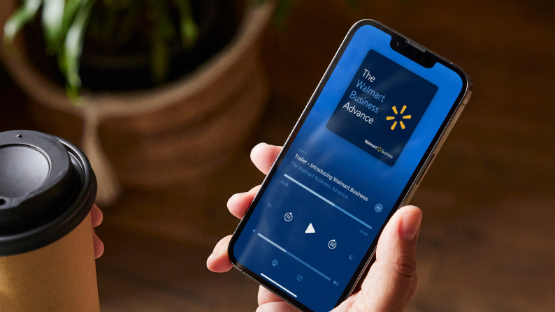 Person holding a cup of coffee and a phone that’s displaying The Walmart Business Advance podcast. 