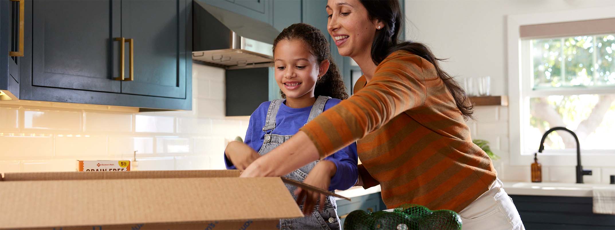 A mother and child unpack a box in the kitchen.