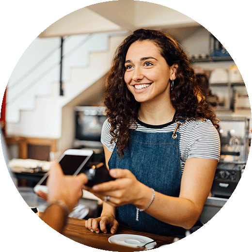 Female shopkeeper with brown hair wearing a denim apron receiving payment through mobile technology in a rustic coffeeshop.